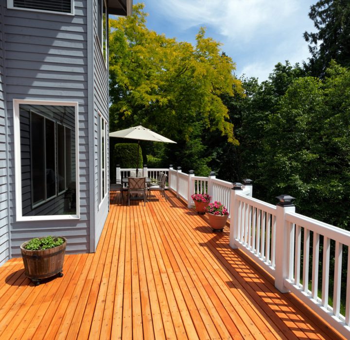 A bright orange wooden deck attached to a gray-sided house, featuring white railings, potted plants, and an umbrella-covered seating area in the distance. The deck is surrounded by lush green trees under a sunny sky.