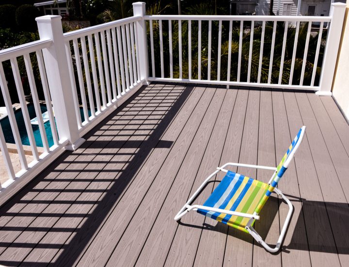 A bright, sunlit deck with light gray composite decking and white railings, featuring a colorful striped lounge chair. The deck overlooks a swimming pool and tropical landscaping, perfect for relaxing by the water.