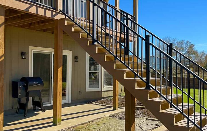 A two-story home with an elevated deck accessed by a wide wooden staircase with black metal railings. The lower level includes a covered patio area with a grill and stone pavers, set against a bright blue sky.