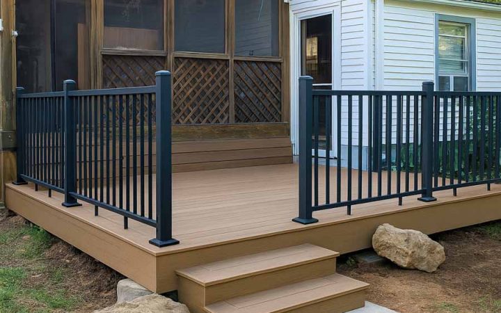 A low-profile backyard deck with a smooth tan-colored surface and black metal railings. The deck features steps leading up from a gravel path, surrounded by rocks and adjacent to a screened-in porch.
