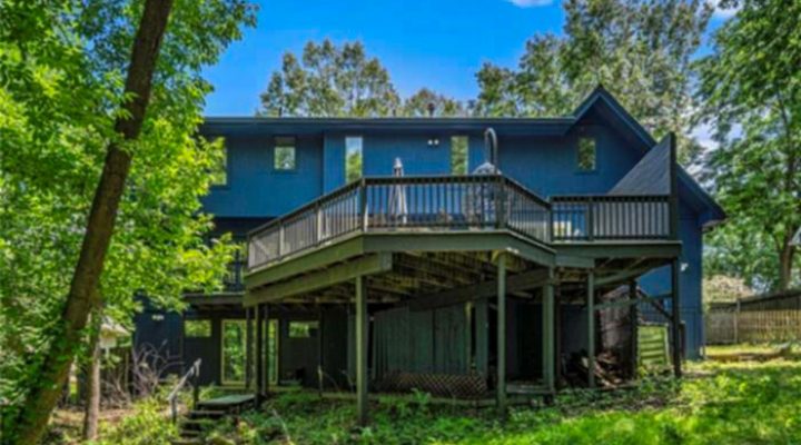 A rustic two-story deck attached to a vibrant blue house, surrounded by dense green trees. The deck features wooden railings and supports, blending into a natural, wooded backyard setting.